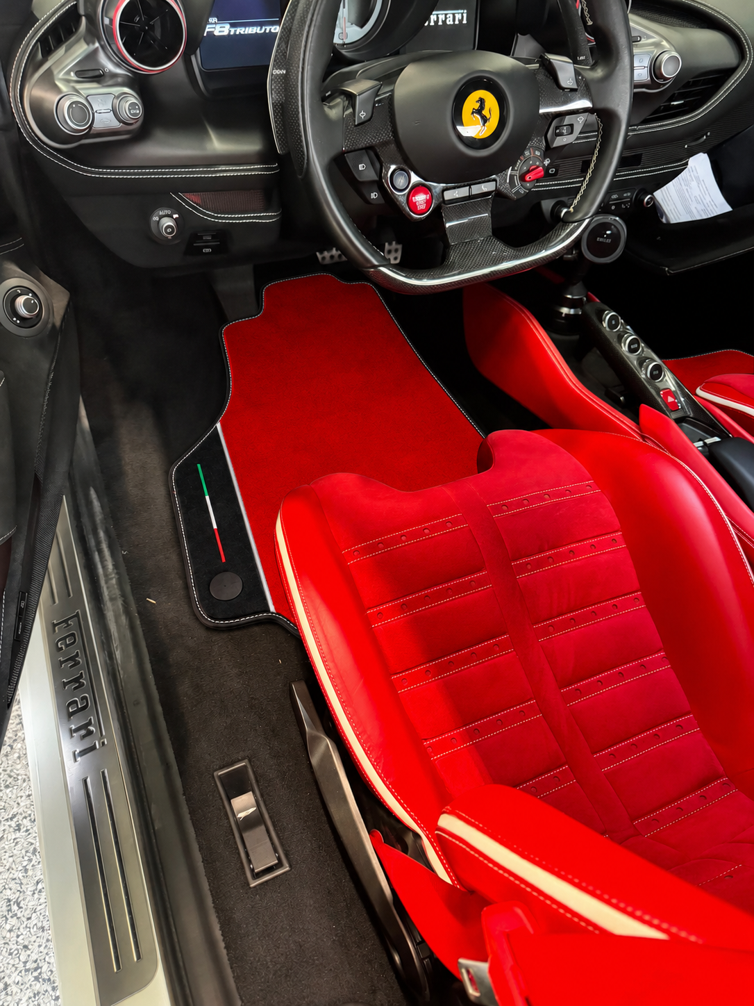 A pair of custom red car floor mats installed in a luxury Ferrari interior, shown alongside the red leather seats and black dashboard.